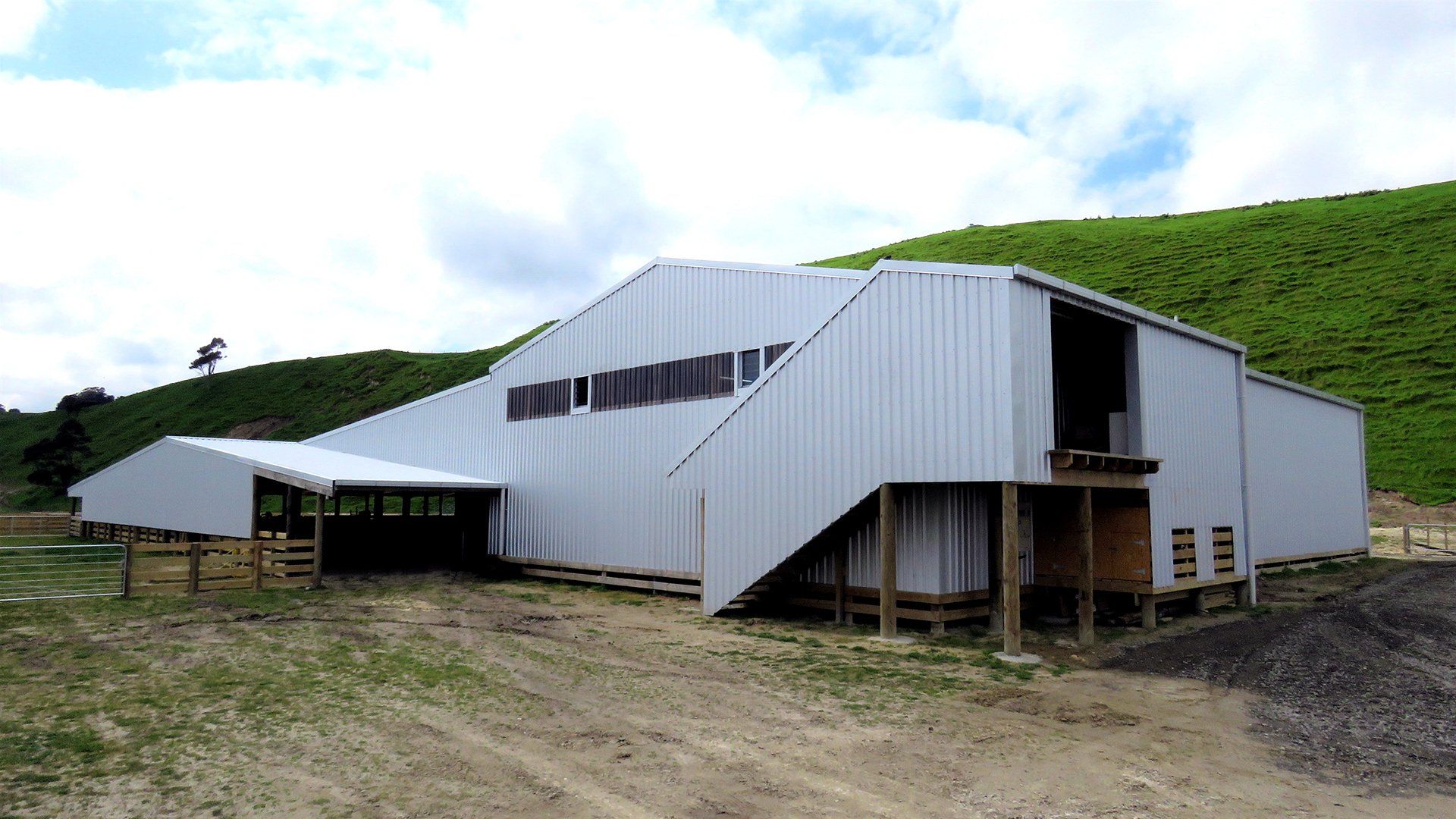 A large metal agricultural shed with a sloped roof stands in a grassy, hilly field under a cloudy sky.