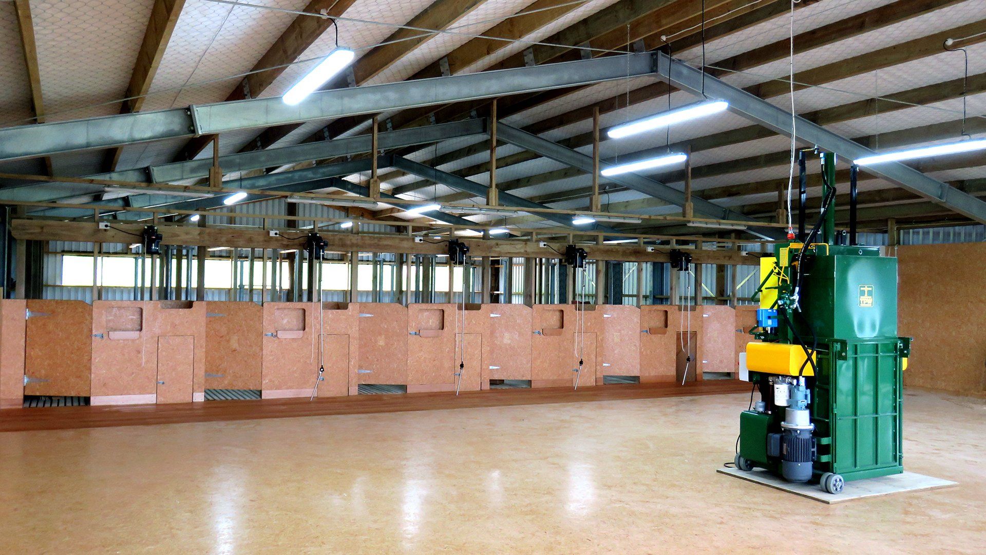 A green industrial waste baler stands on a clean floor inside a large, open-plan warehouse with wooden partitioned stalls.