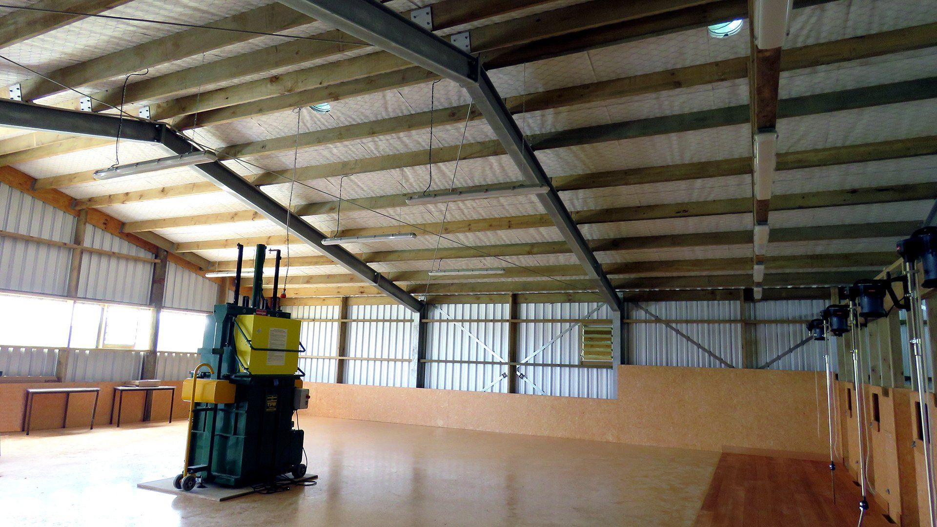 A large, open warehouse featuring a high wooden-beam ceiling, light walls, and a green industrial machine in the center.