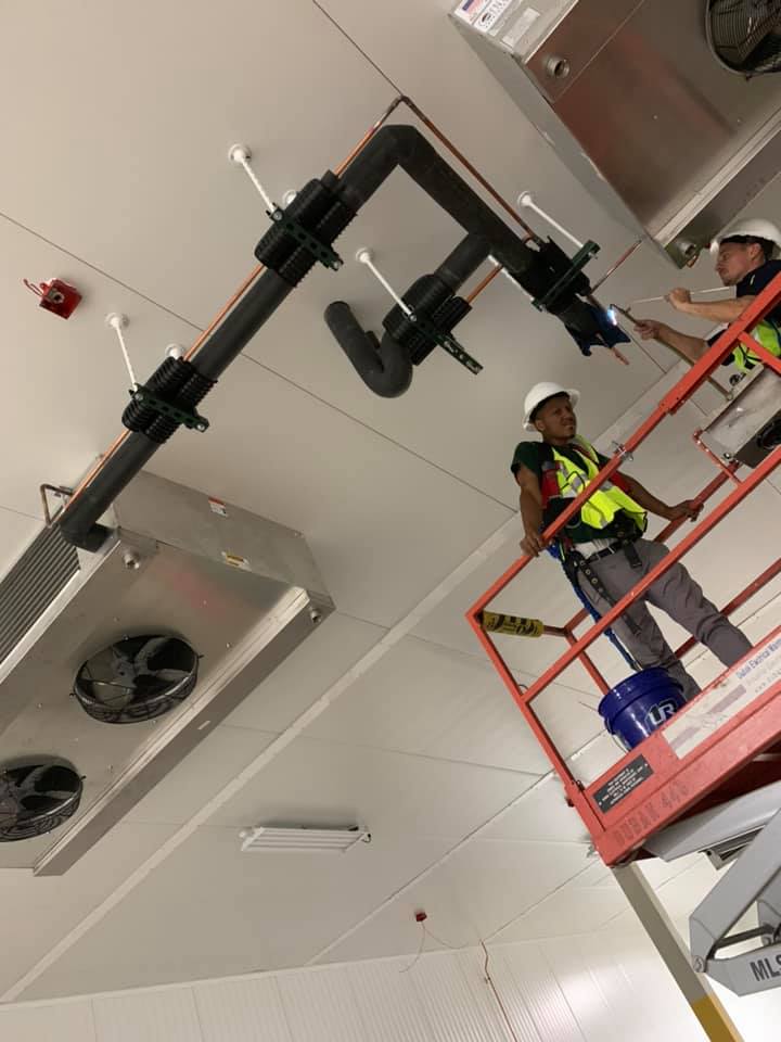 Two workers on a lift installing plumbing on a white ceiling. Large black pipe is attached with clamps.
