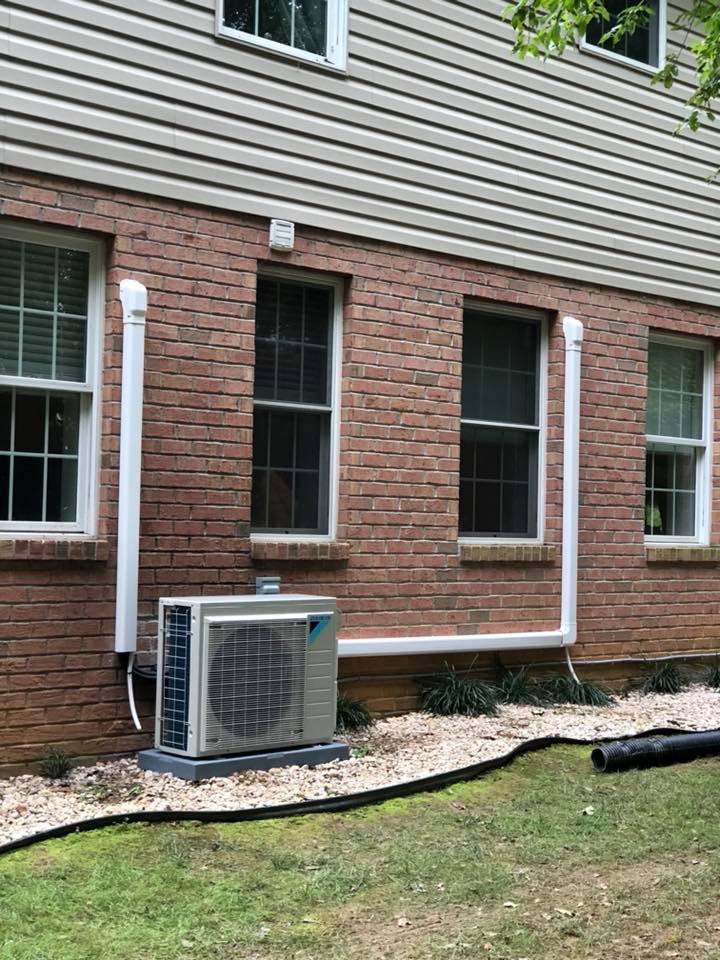 Exterior view of brick building with a heat pump unit. White pipes ascend the wall. Green grass and ground cover.