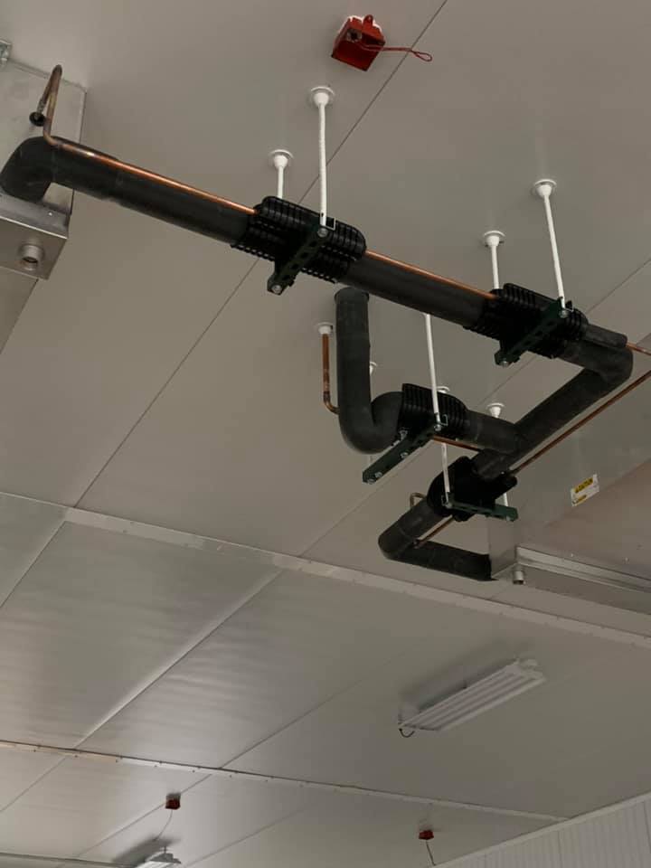 Black pipes suspended from a white ceiling with supporting rods and black clamps, near lights and a red box.