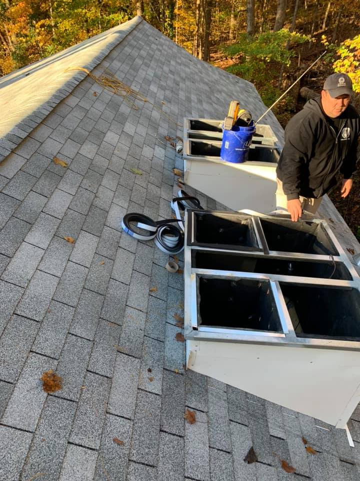 Person on a roof working near rectangular skylights. Gray shingle roof, blue bucket, fall foliage.