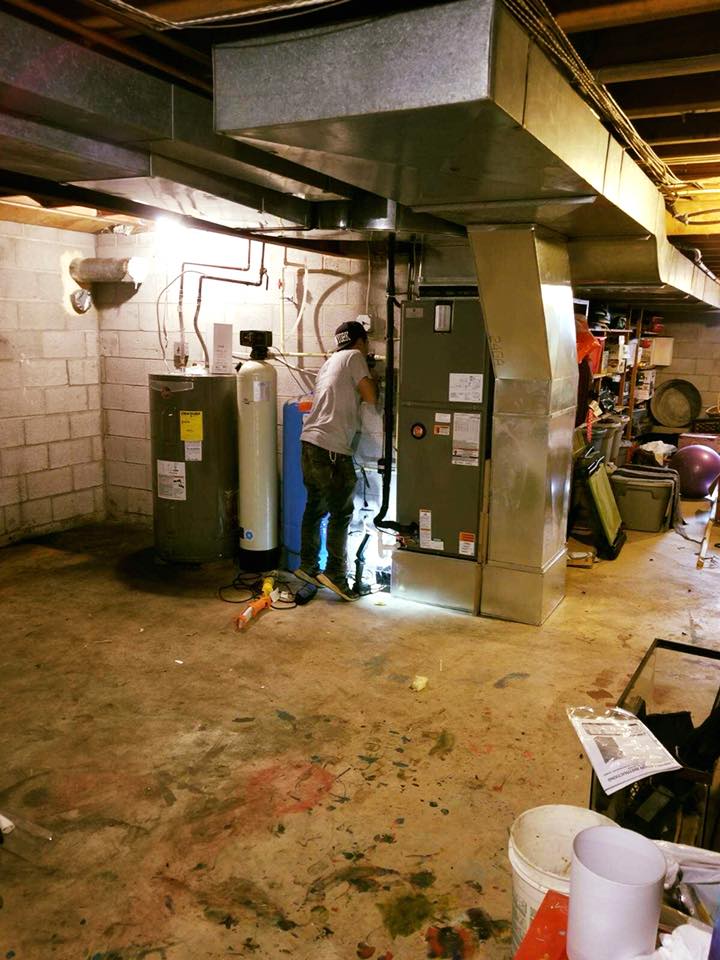 Person working on HVAC system in a basement; water heater and water softener visible.