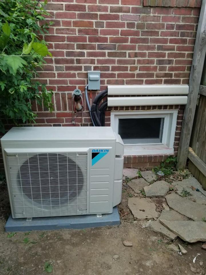 Daikin HVAC unit outside brick wall, near a window and wooden fence.