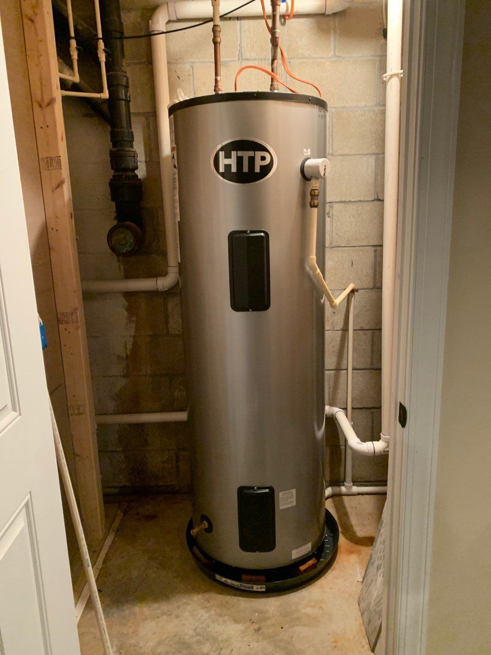Silver HTP water heater in a utility closet with pipes and concrete block walls.
