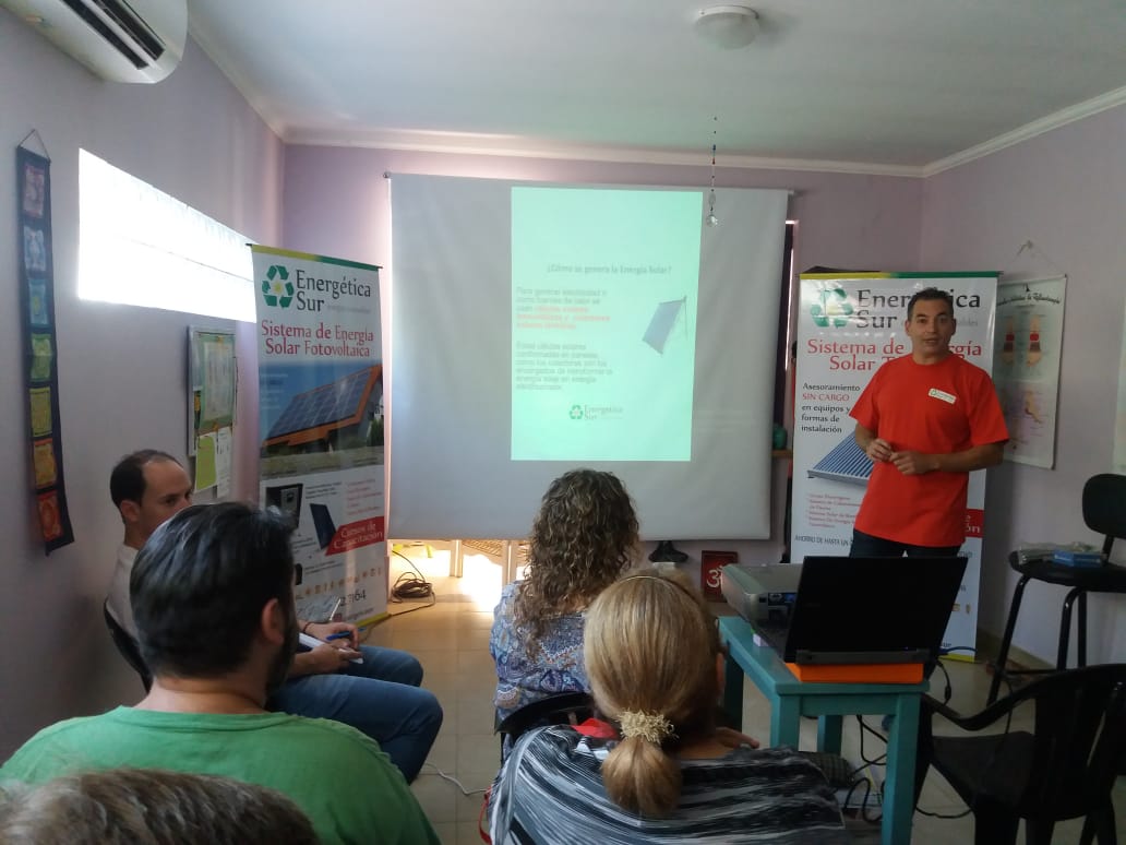 Presentador con camisa naranja hablando a un público sentado durante una presentación proyectada en una sala pequeña.