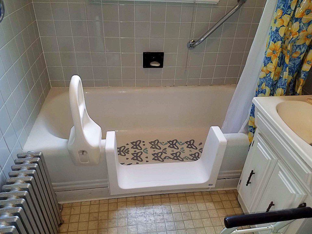 Step-in bathtub with built-in seat in a tiled bathroom, next to a radiator and sink cabinet.