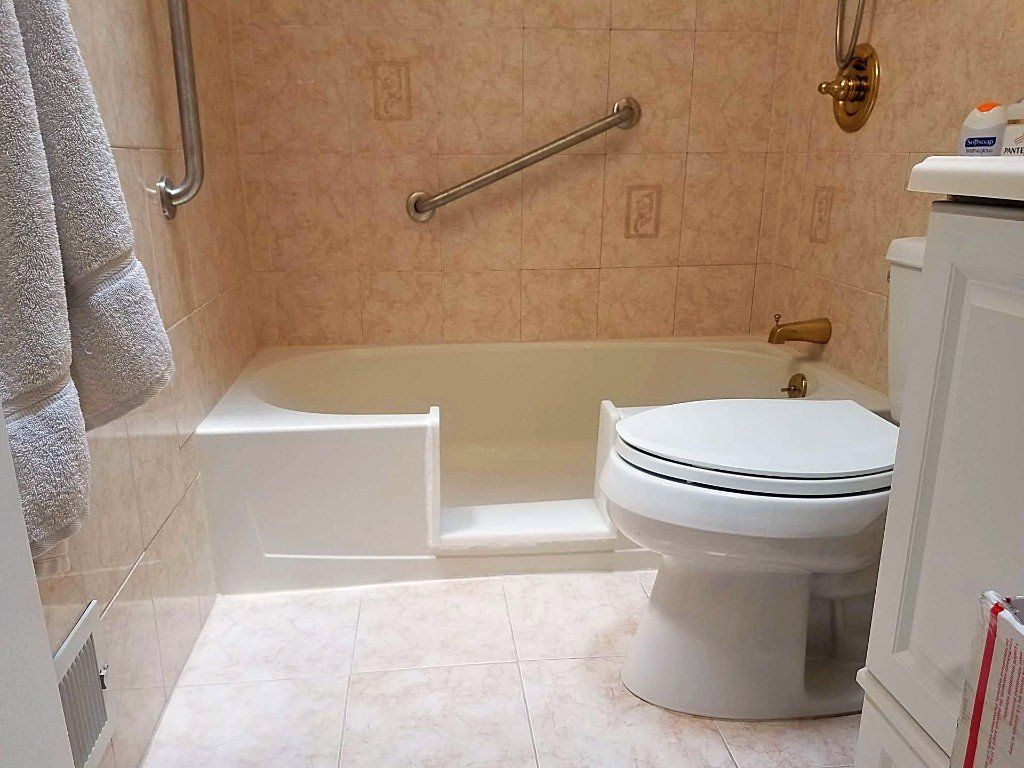 Small beige tiled bathroom with a step-in tub, raised shower step, and toilet beside a white cabinet.