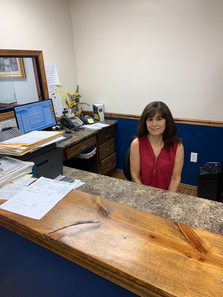 Woman sitting behind a reception desk in an office, with papers, computer monitors, and a mirror on the wall.