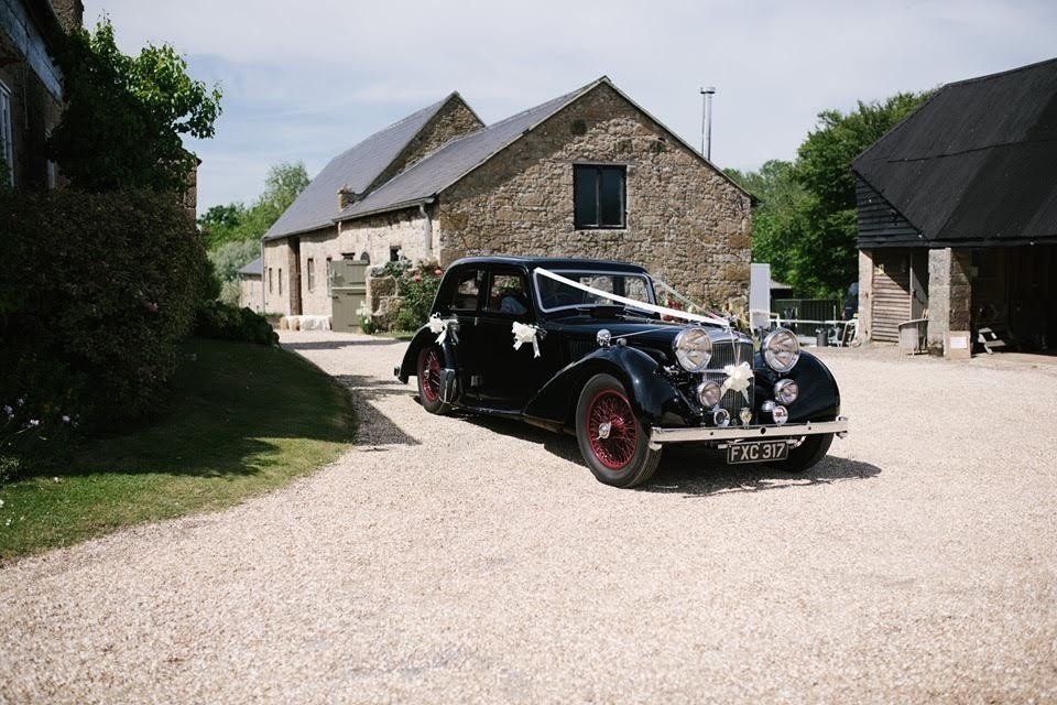 A black car is parked on a gravel road in front of a stone building.