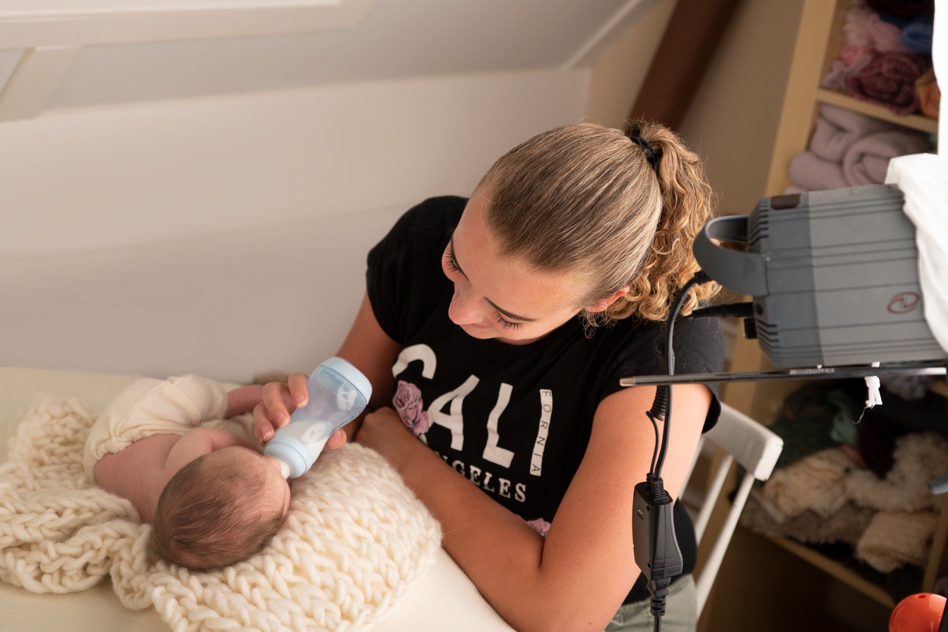 Een persoon geeft een baby de fles in een studio-omgeving. De persoon draagt ​​een zwart shirt en kijkt naar de baby.
