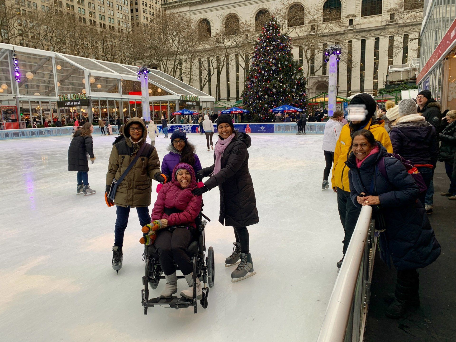 It's a festive scenery in the background with the famous Christmas Tree. The family, incl wheelchair user, is on ice smiling.