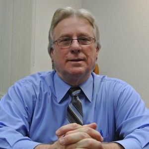 Man in a blue shirt and striped tie, seated with hands clasped, looking at the camera.