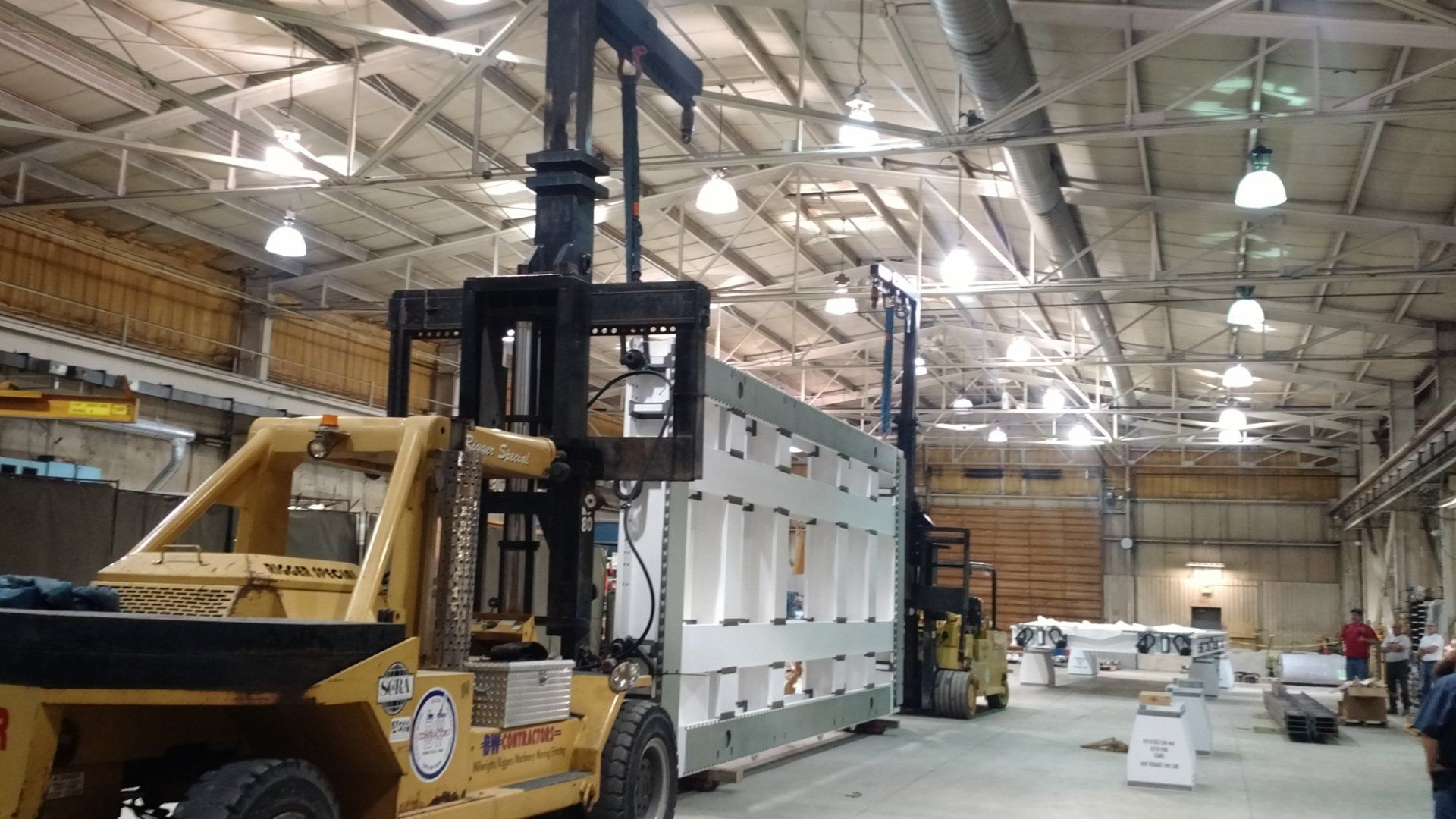 Forklift in a warehouse lifting a white wall panel beside a yellow loader and work trucks.