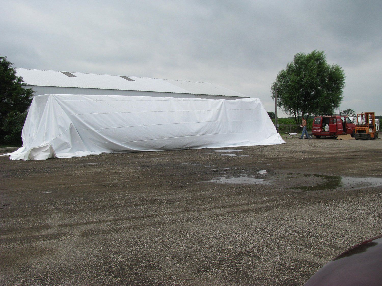 Large white tarp-covered structure on a gravel lot, with a tree and equipment in the background.