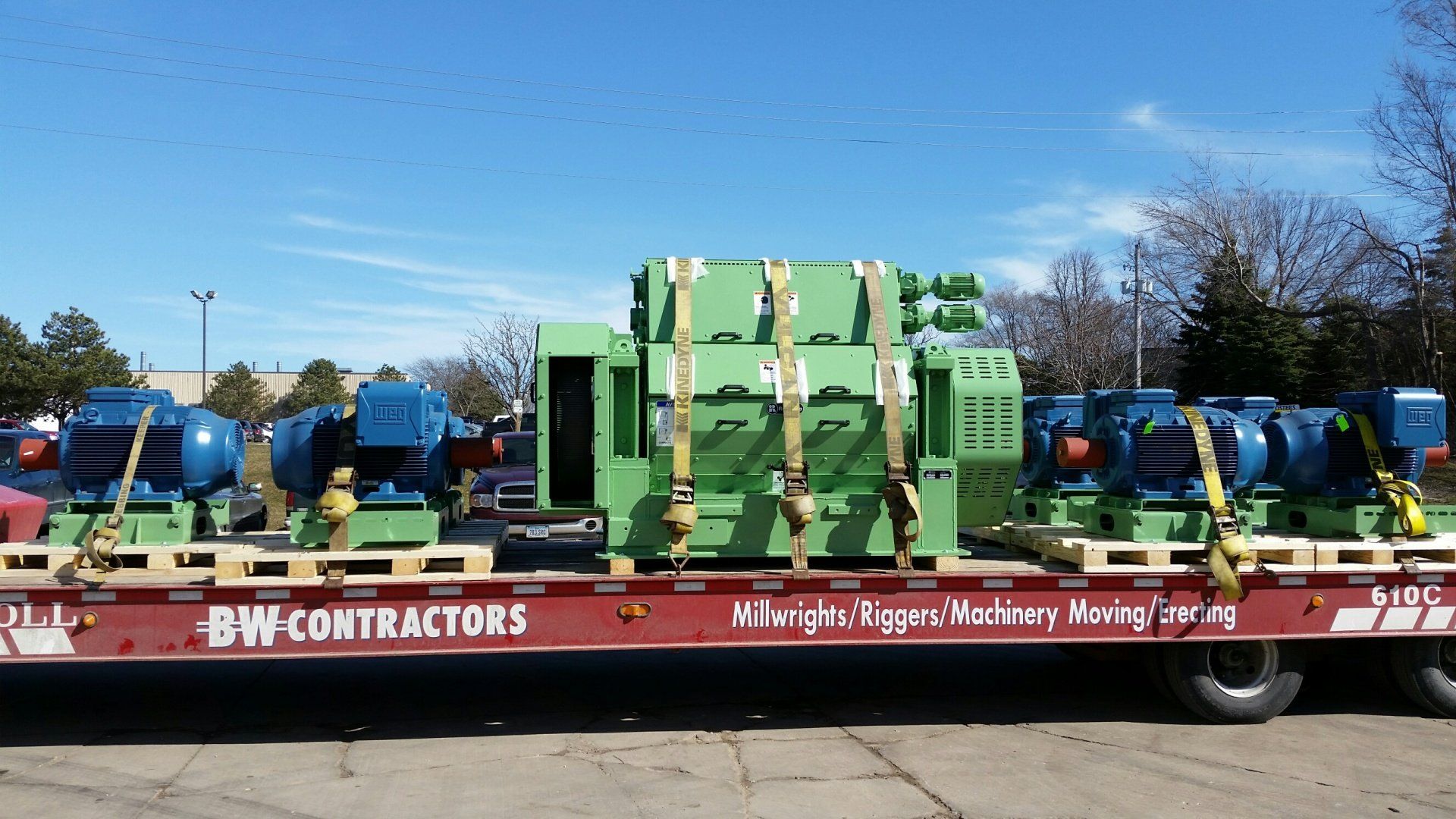 BW Contractors flatbed trailer carrying green industrial machinery and blue equipment outdoors
