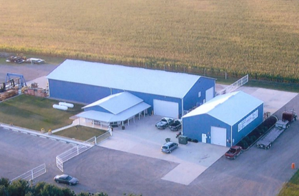 Aerial view of two blue industrial buildings with a parking lot and surrounding fields.