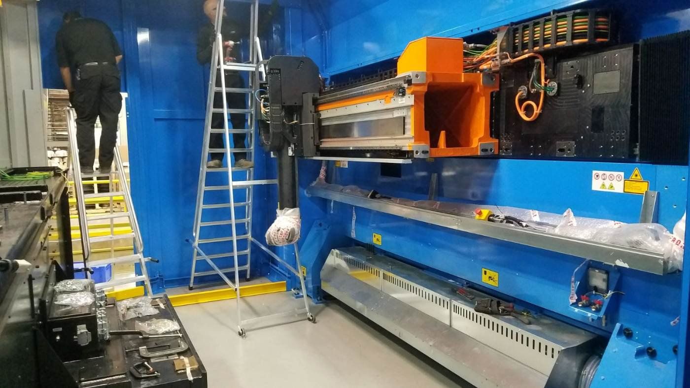 Blue industrial machine with orange carriage and ladder, in a factory-like workspace.