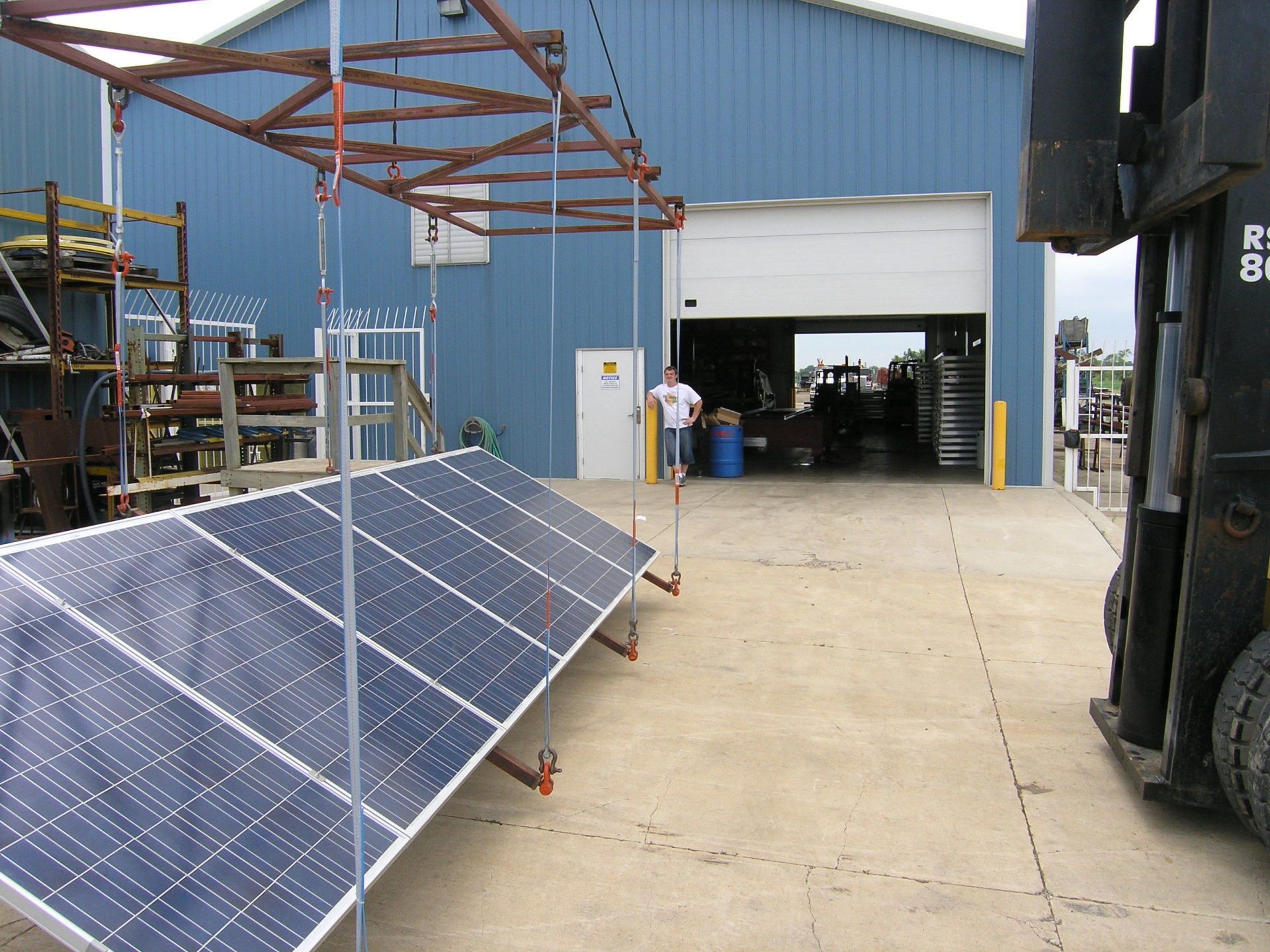 Double Crane Lifting Heavy Machine — Cedar Falls, IA — BW Contractors Inc. Solar panels on a concrete lot beside a blue warehouse with an open garage door and forklift