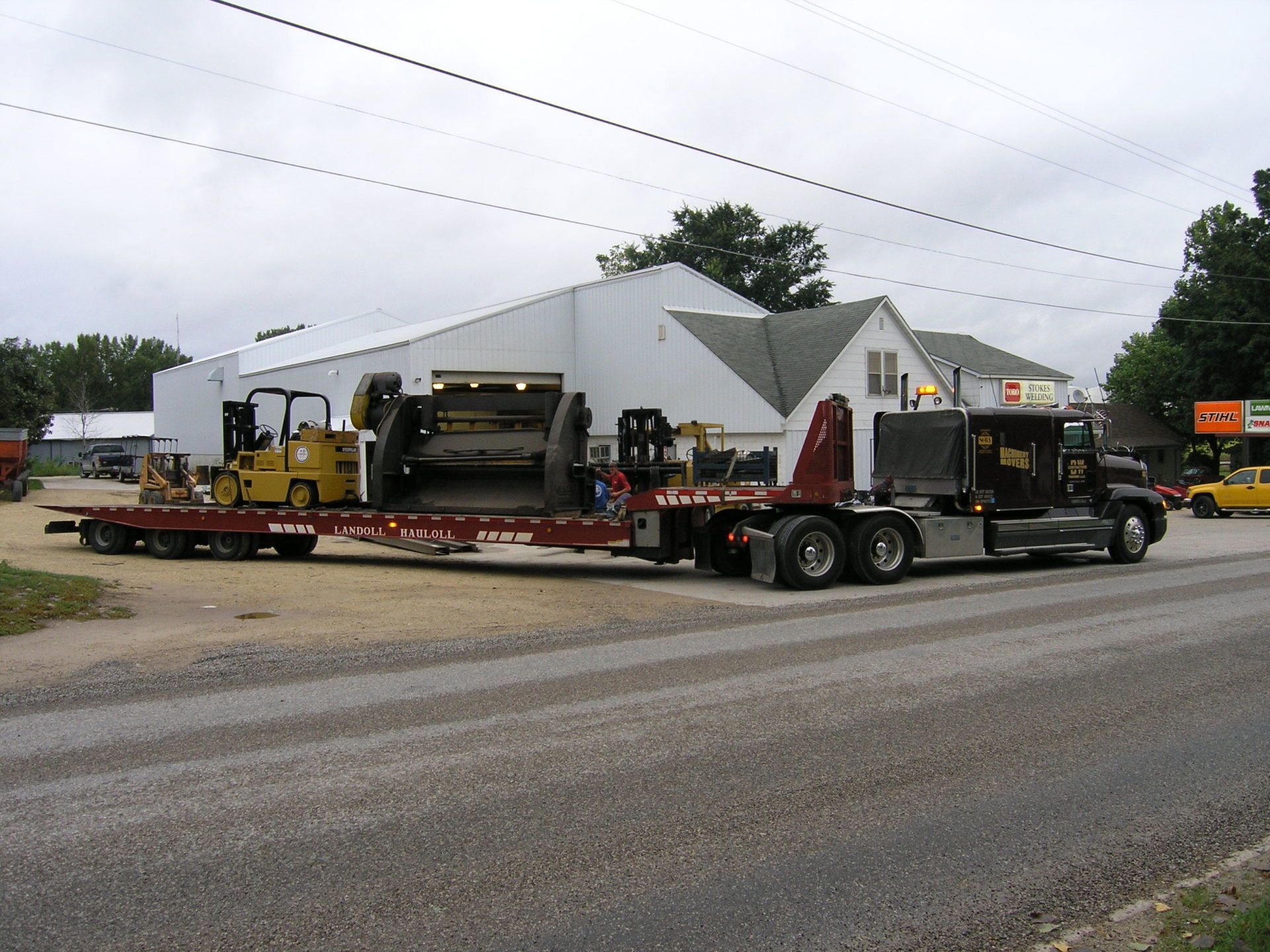 Heavy-duty tow truck hauling yellow construction equipment on a flatbed by a roadside.