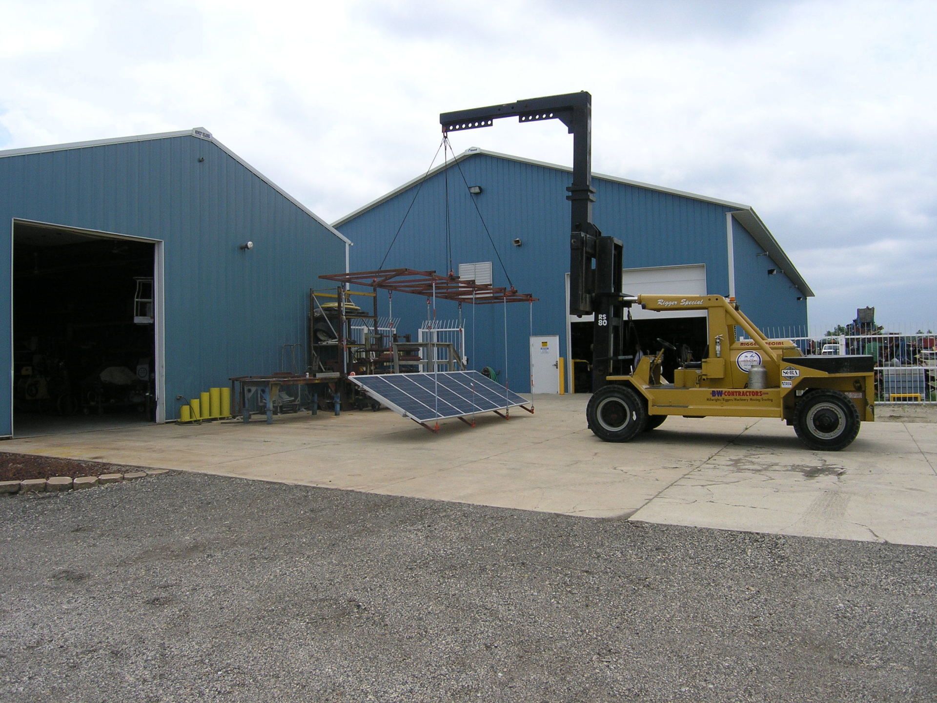 Blue industrial buildings with a yellow forklift and a crane in a gravel yard