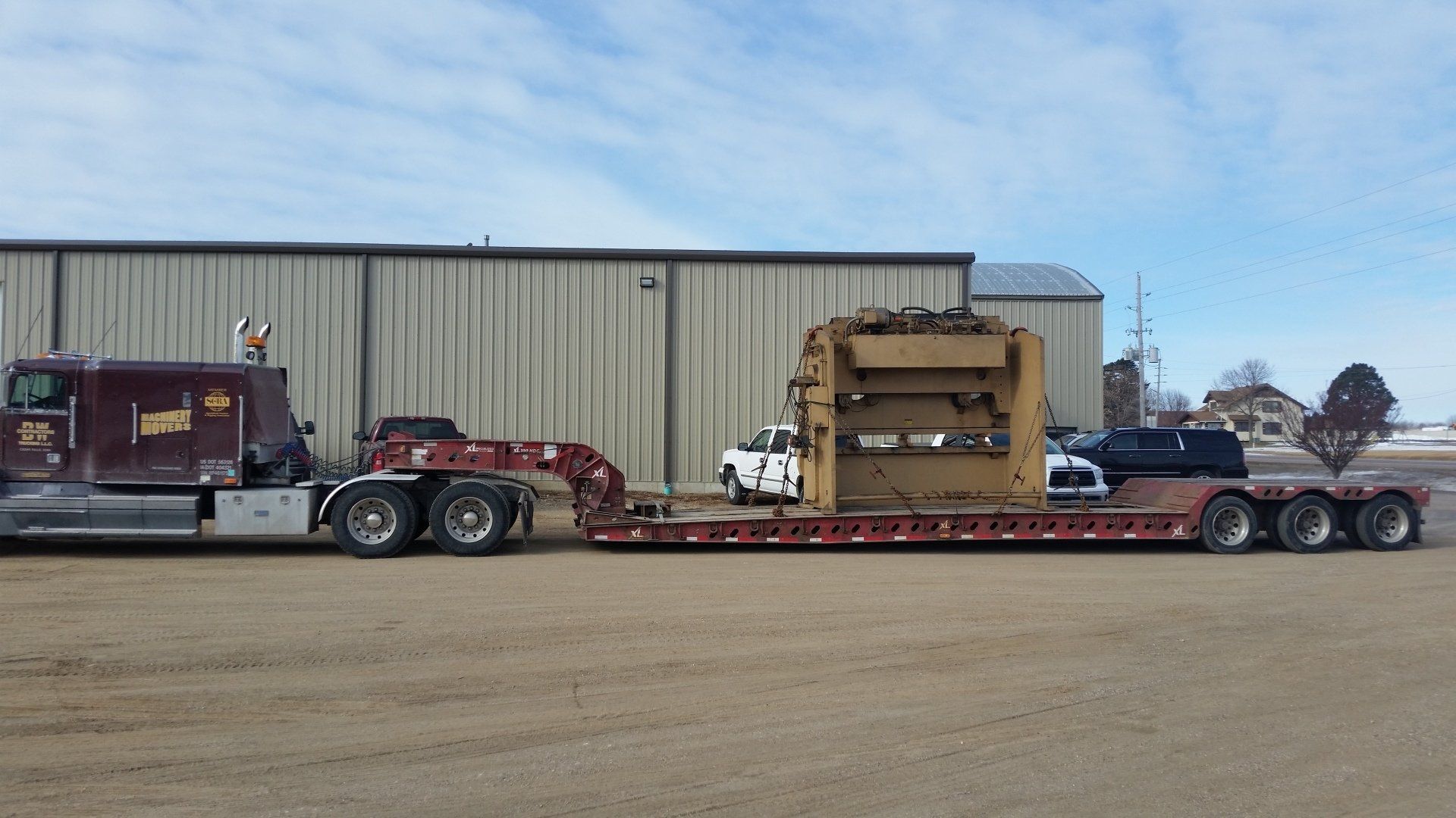 Heavy machinery on a long flatbed trailer parked outside a warehouse under a clear sky.