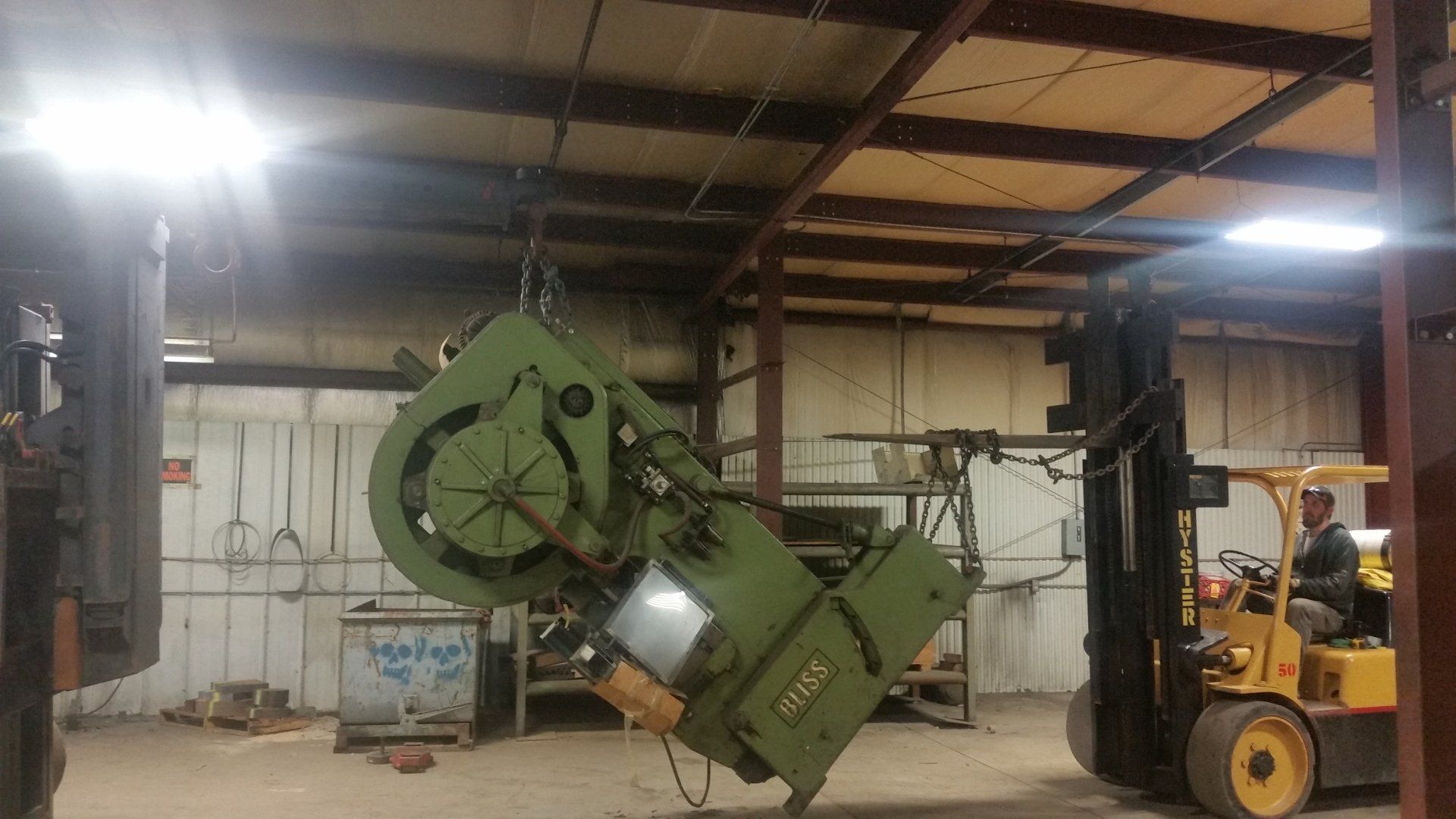 Green industrial machine being lifted by a yellow forklift inside a warehouse.