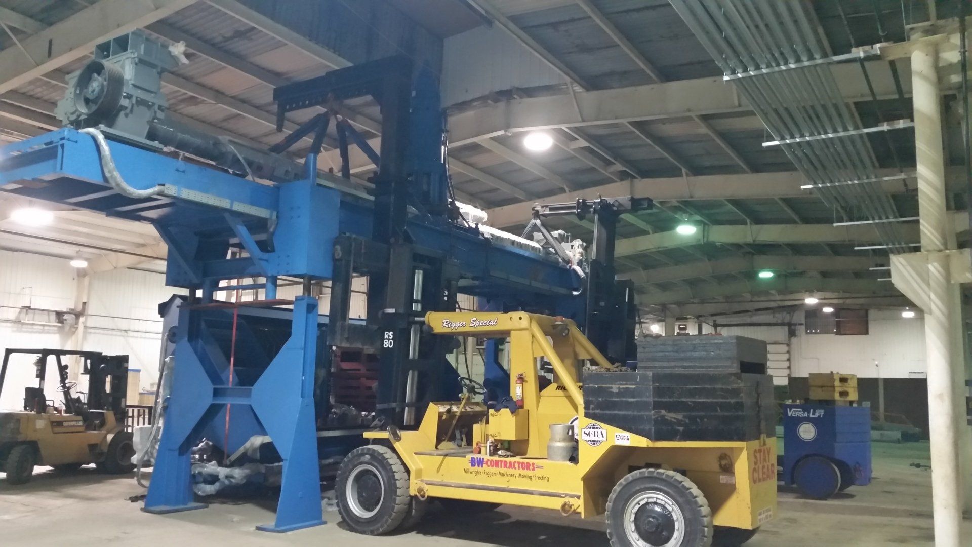 Yellow forklift beside a large blue industrial machine inside a warehouse-like factory.