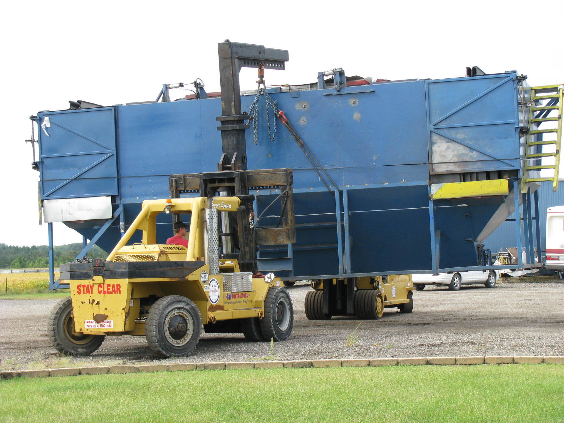 Yellow forklift moving a large blue industrial tank on a gravel lot