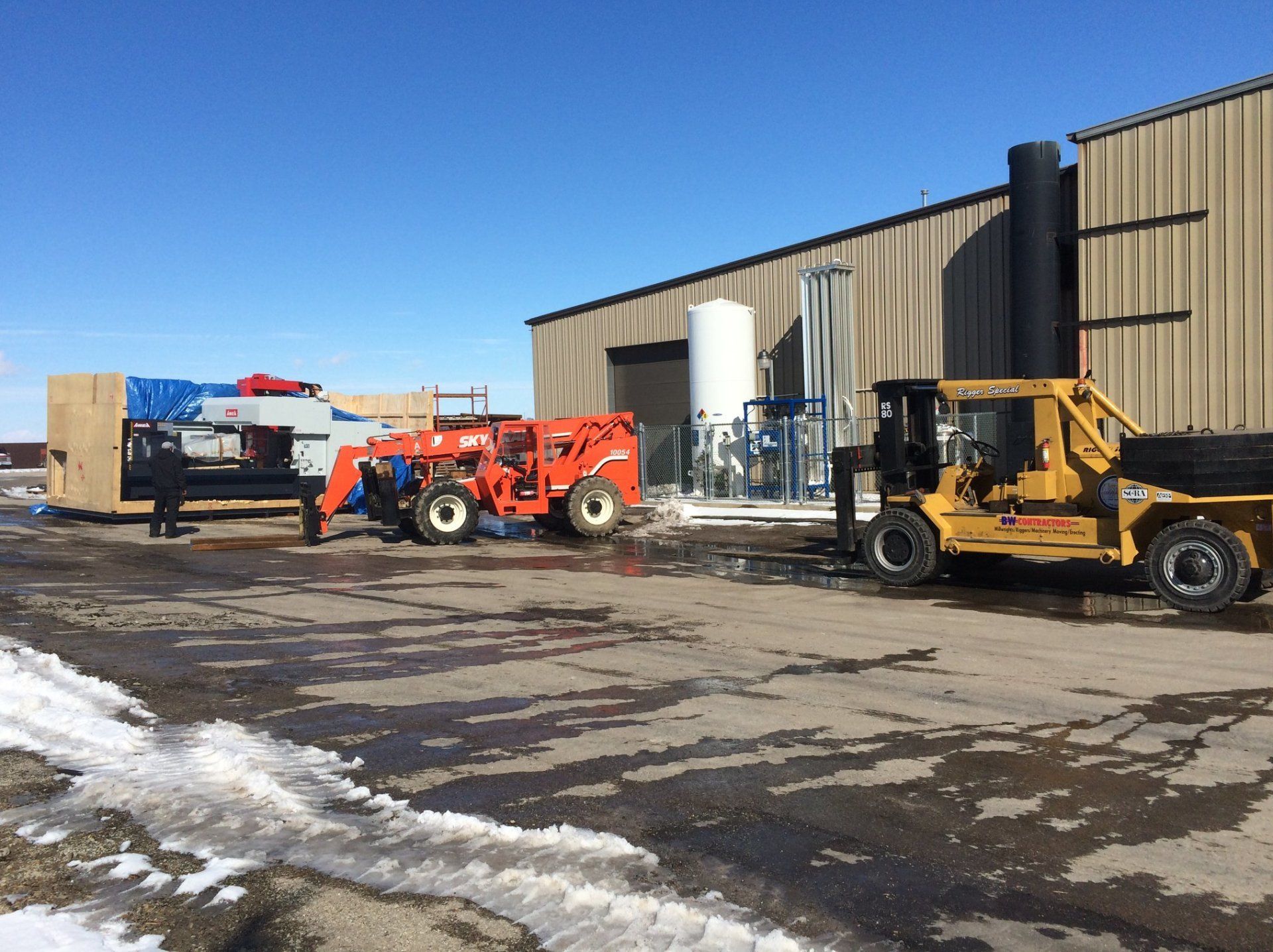 Construction equipment parked beside a warehouse on a snowy industrial lot