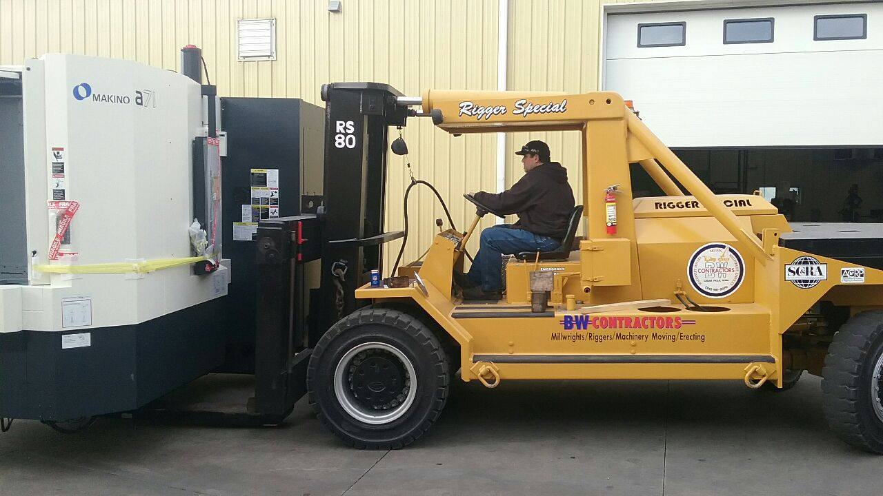 Tow tractor towing a large industrial machine in a warehouse, with a seated operator inside the cab.