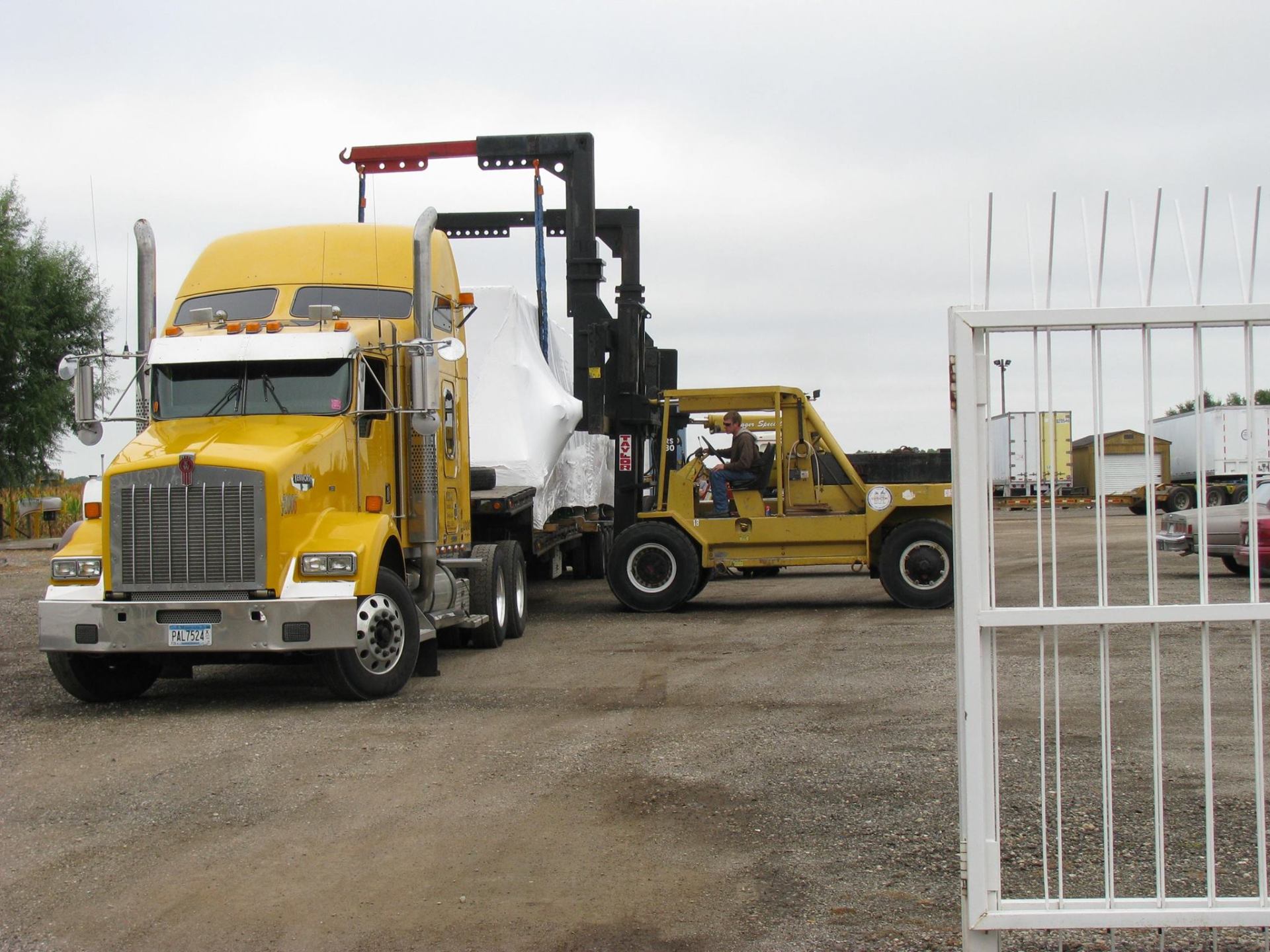 Yellow semi-truck loading a white wrapped machine onto a flatbed at an industrial yard