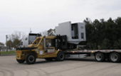 Yellow forklift towing a flatbed trailer carrying a white modular building on a lot