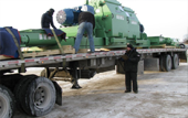 Workers loading a large green machine onto a flatbed trailer in a snowy outdoor setting