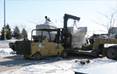 Yellow tow truck lifting a gray van in a snowy lot with a car parked beside it