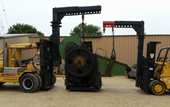 Two yellow forklifts lifting a large black industrial machine outdoors