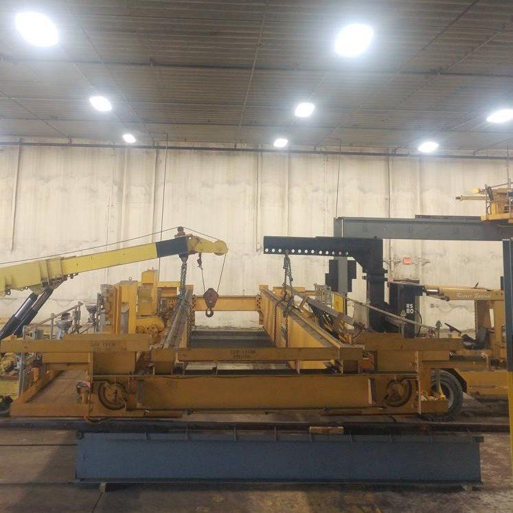 Yellow industrial machine in a warehouse, with cranes and steel beams under bright ceiling lights.