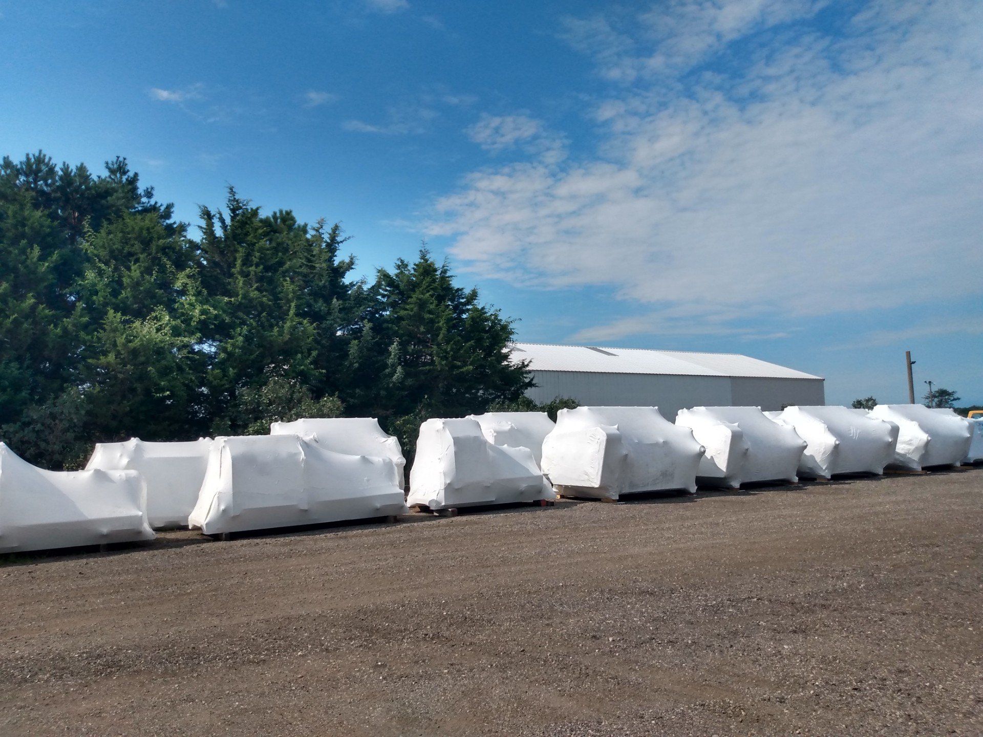 White wrapped bales lined beside a gravel lot under a blue sky, with trees and a low building in the background