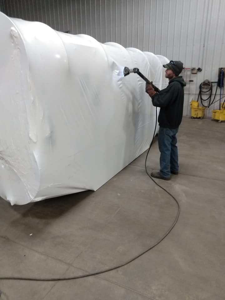 Worker spray-paints a large white wrapped object in an industrial warehouse.
