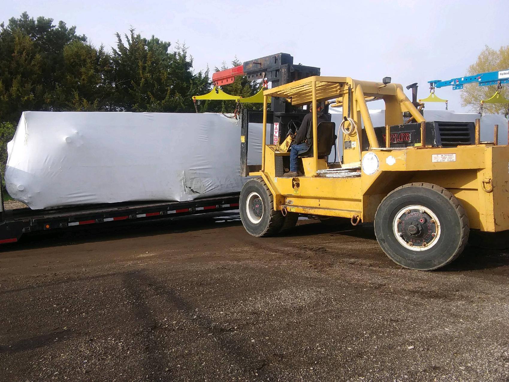 Yellow forklift moving a large wrapped load on a trailer in an outdoor yard