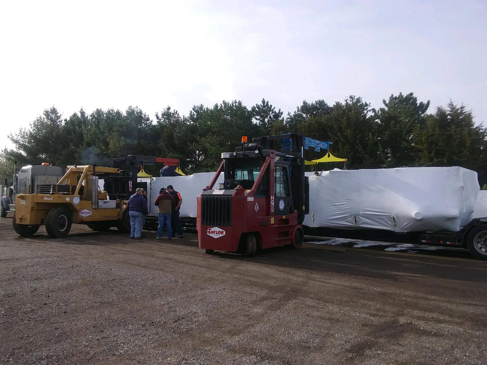 Forklifts loading a large wrapped white bundle in an outdoor gravel yard