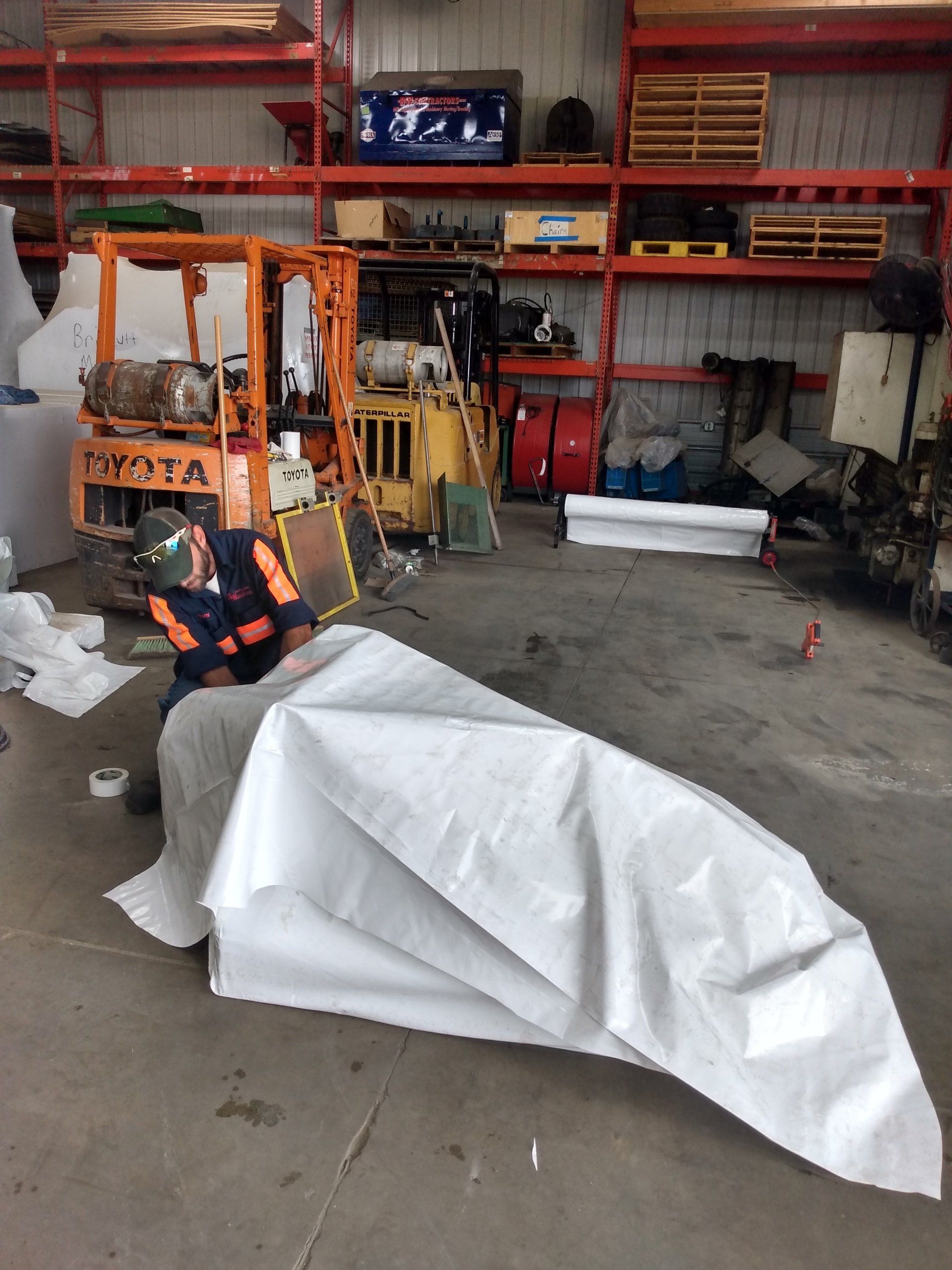Person in an orange jacket works beside a tarp-covered object in a warehouse garage