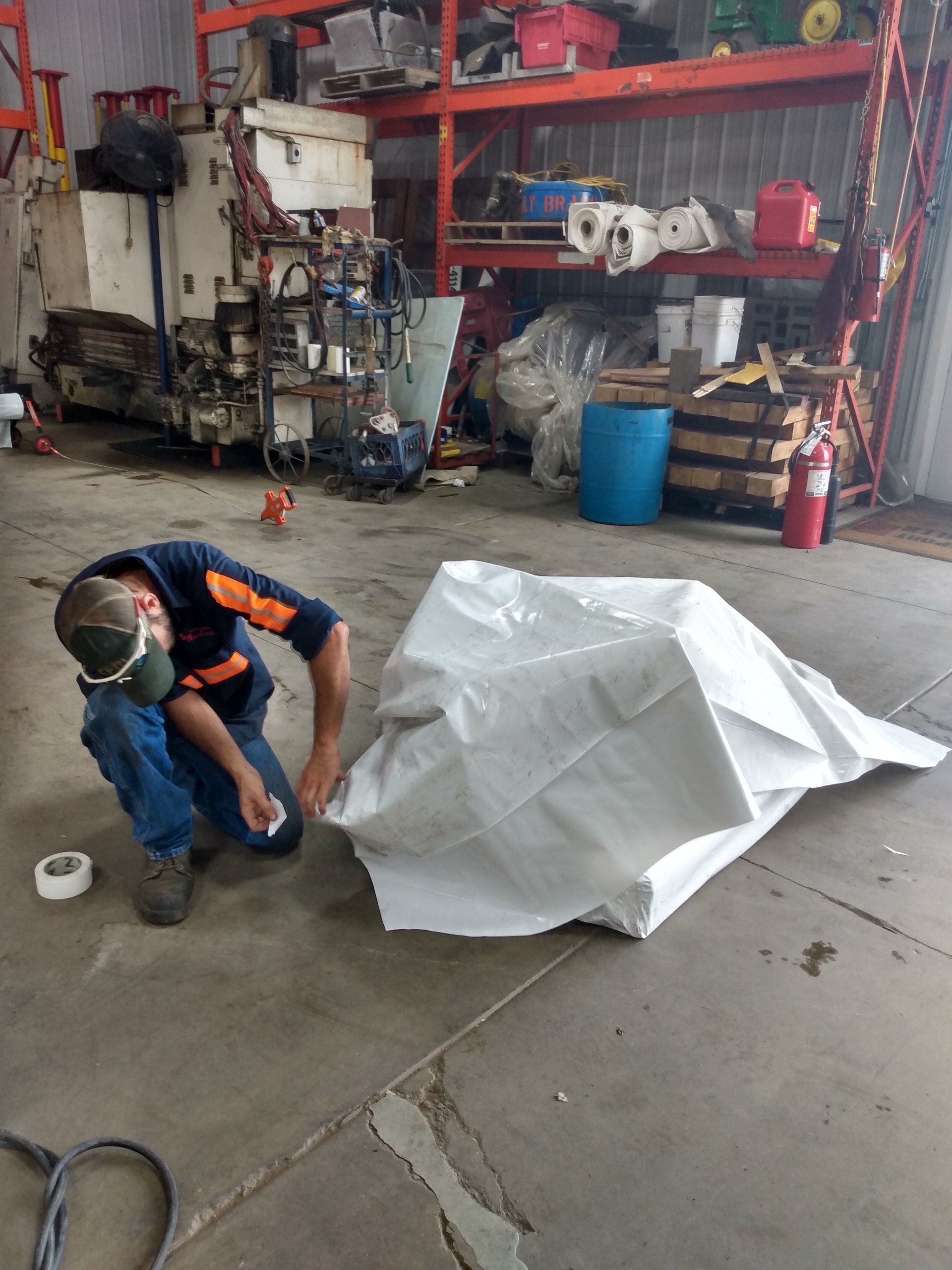 Worker kneels on a concrete floor in a workshop beside a large white covered object and a roll of tape.