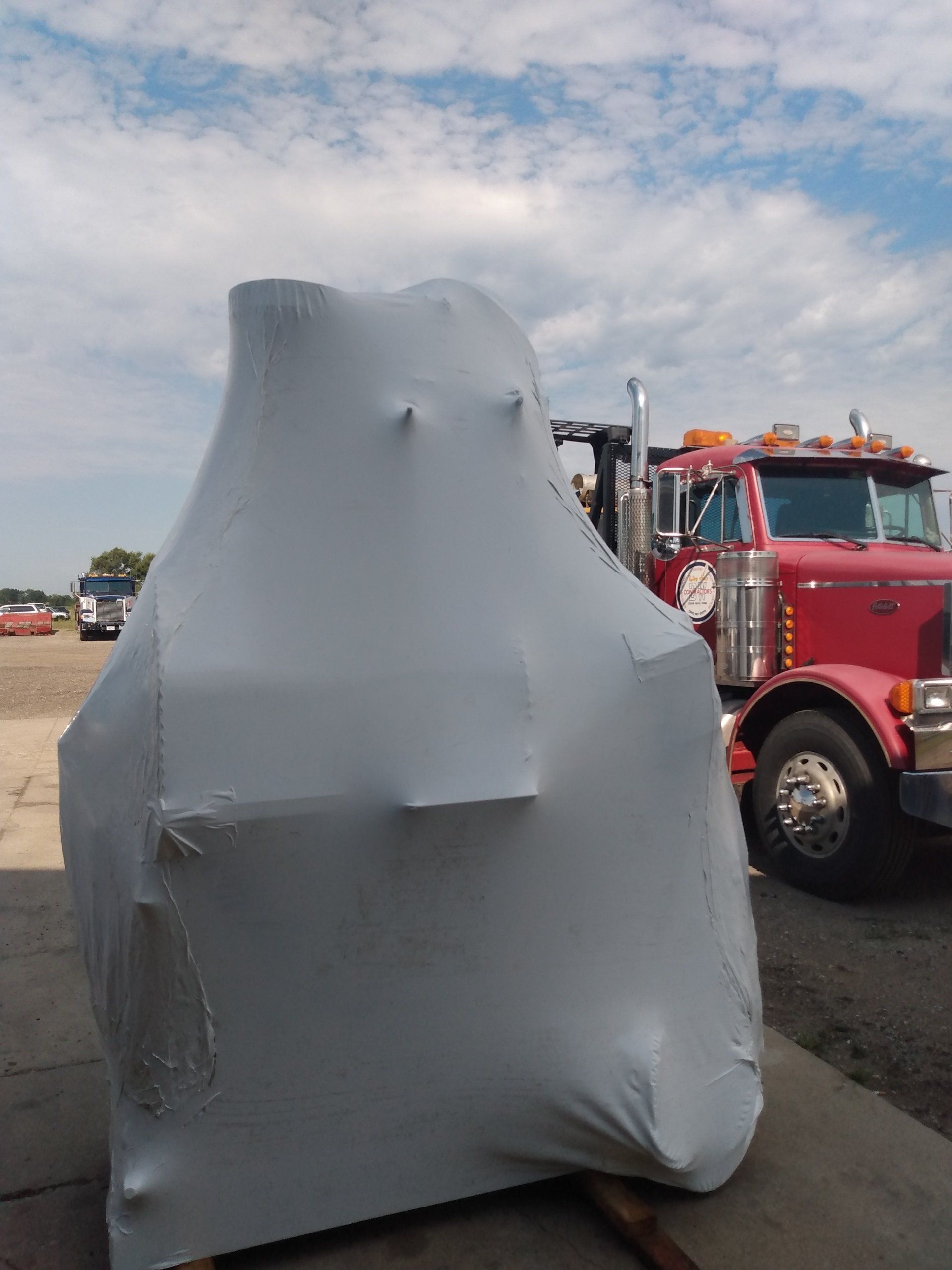 Gray object cover in foreground with a red truck parked behind it on a dirt lot under a cloudy sky