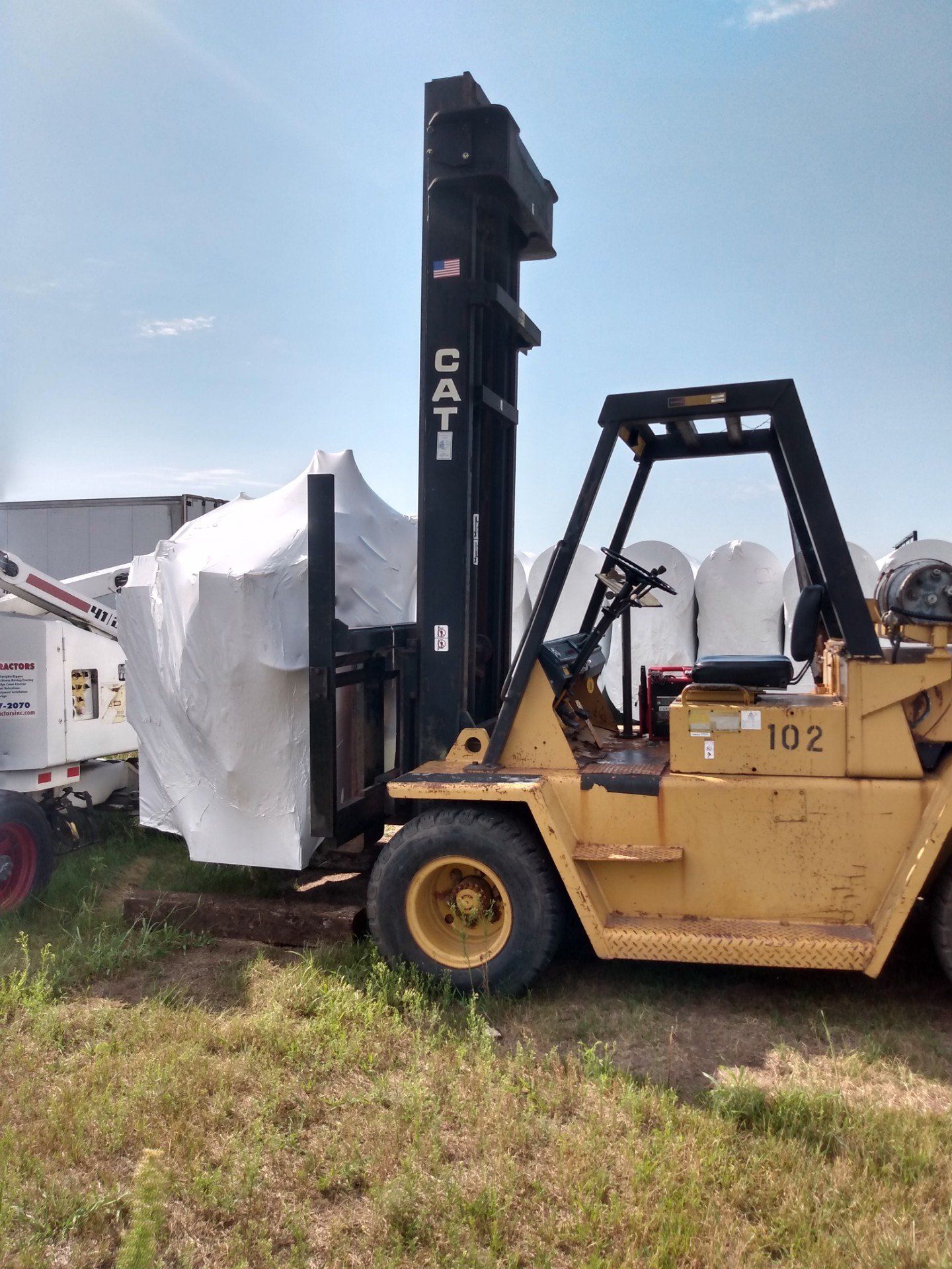 Yellow CAT forklift beside a white tarp-covered load in a grassy outdoor lot