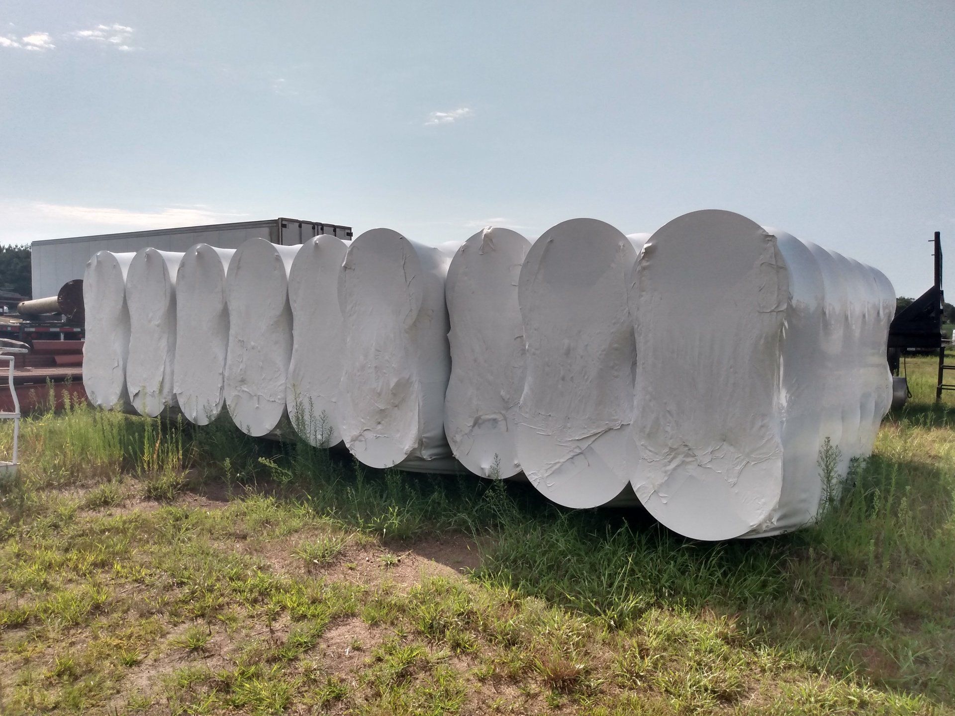 White plastic-wrapped hay bales stacked in a row in a grassy field under a cloudy sky