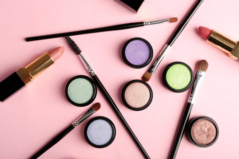 Prodotti per il trucco, tra cui rossetto, ombretto e pennelli, disposti su una superficie rosa.