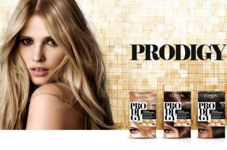 Modella bionda con scatole di tinture per capelli L'Oréal Prodigy su uno sfondo dorato decorato.