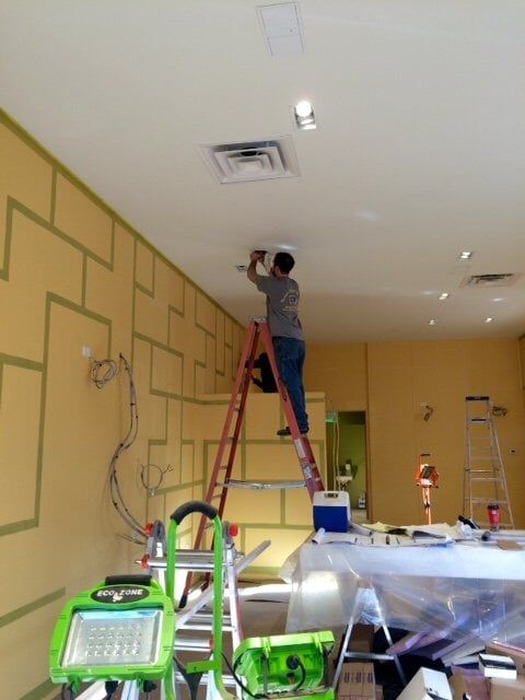 Person standing on a red ladder painting a beige wall in a room under renovation.